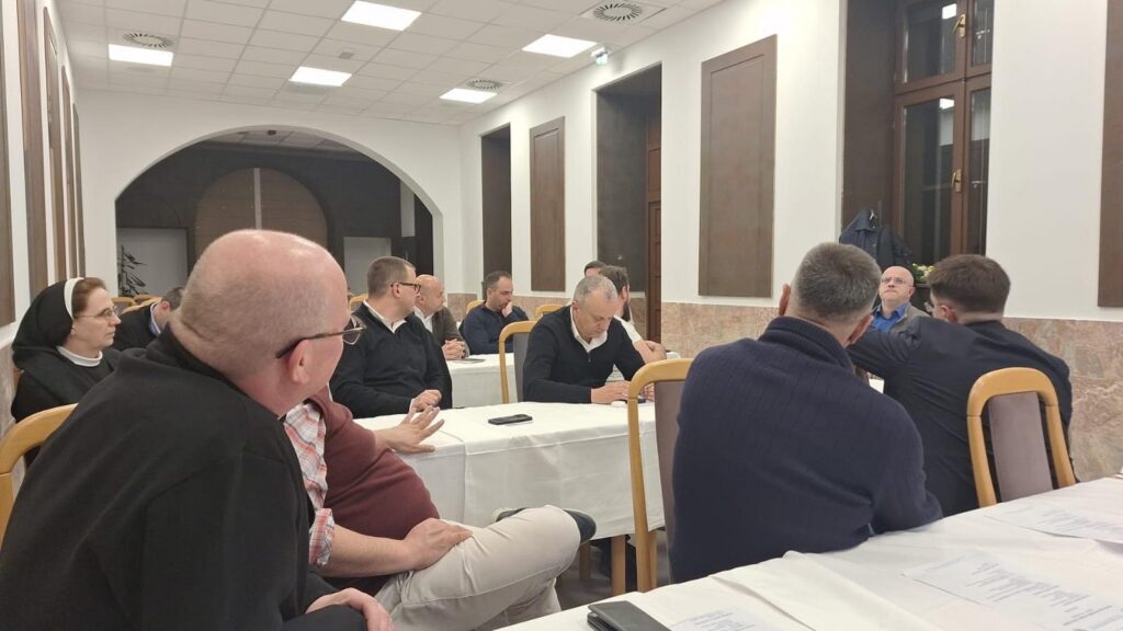 Group of adults seated around a long conference table in a well-lit room, reviewing documents and talking together.] ,