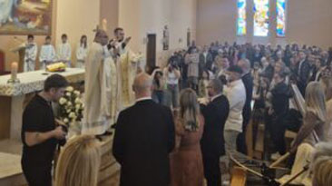 Inside a church during a ceremony with priests in white vestments at the altar as congregants stand nearby, stained glass visible in the background.