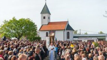A large crowd gathers outside a small church with a red roof and steeple during a daytime event.