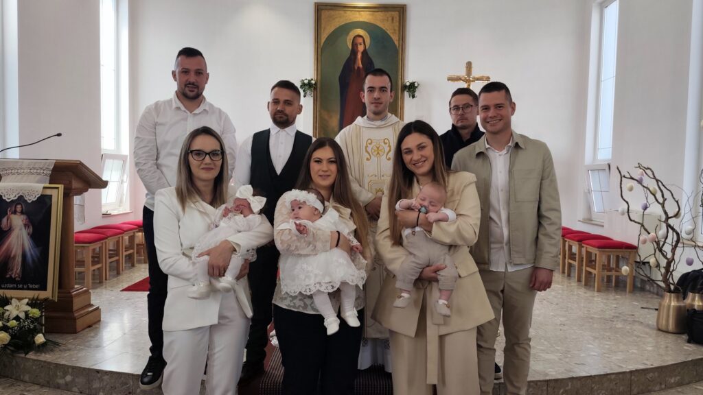 Family and friends pose for a baptism photo in a church, with the priest at the altar behind them.
