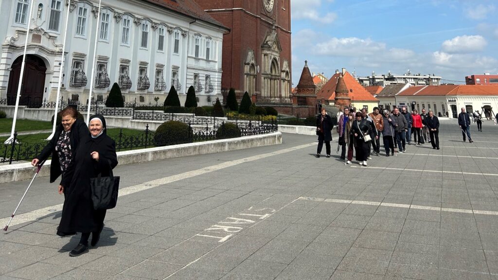 A nun in a black habit and a companion lead a line of pedestrians across a wide plaza beside a historic red-brick church and pale ornate buildings on a sunny day.
