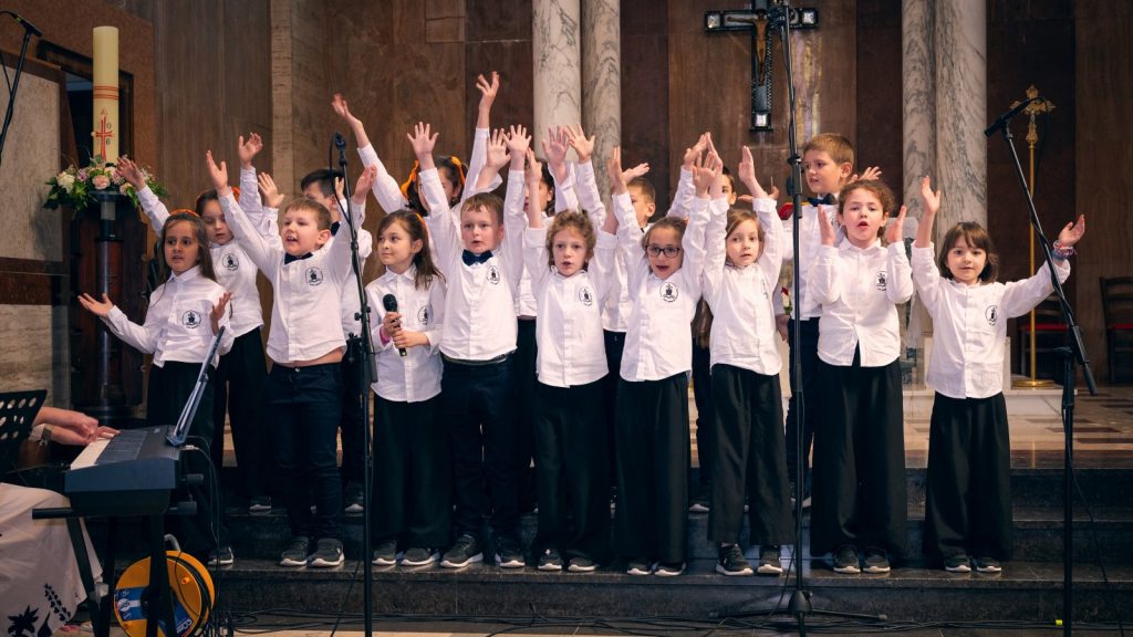 Children’s choir of young students on a church stage, raising their hands as they sing, with a pianist nearby and a crucifix in the background.