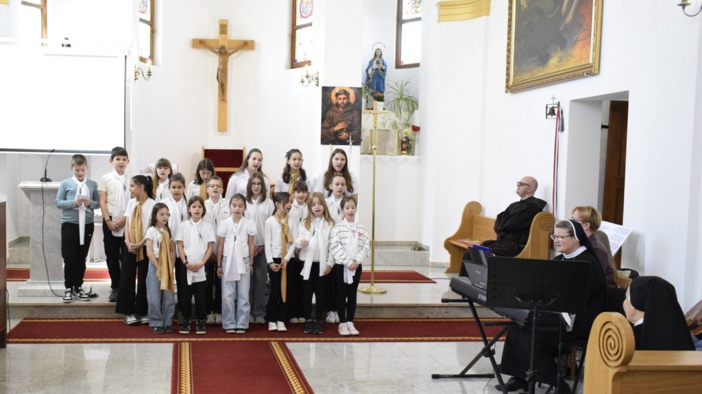 Children choir sings in a church, standing on red-carpeted steps near the altar with a pianist nun and others nearby.