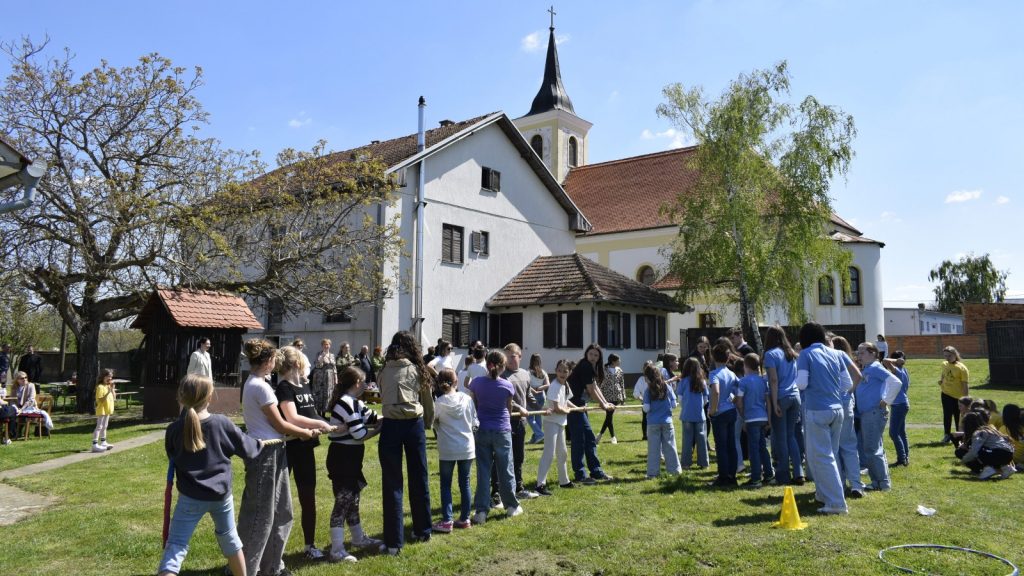 Children and adults outside a white church building, lined up and holding a rope for a group game on a sunny day