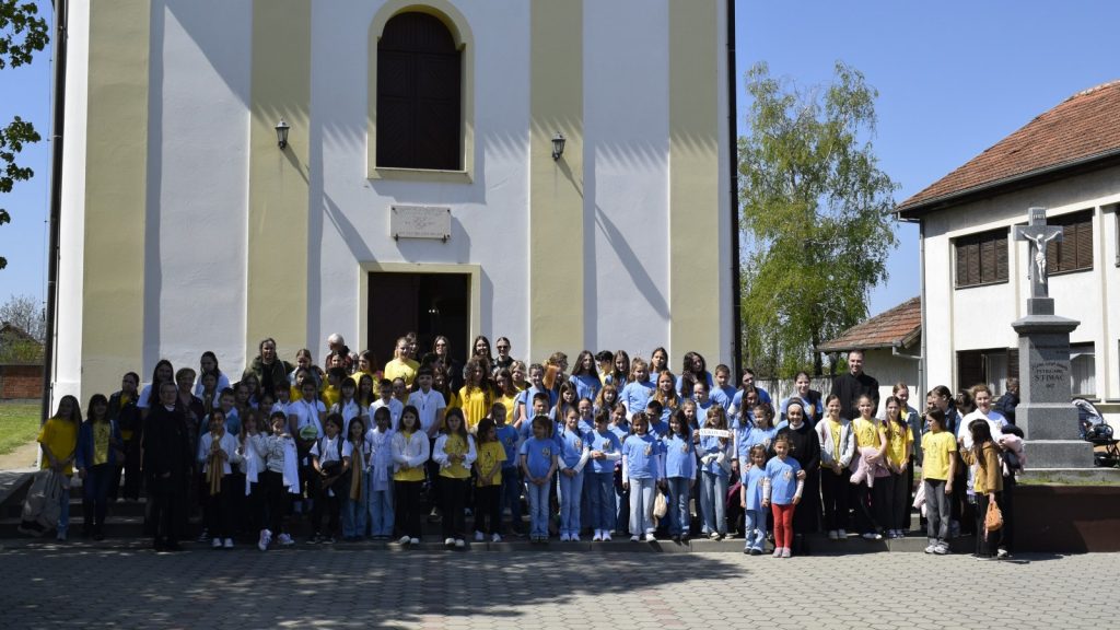 Large group of children and adults posing in front of a pale yellow and white church façade with a tall arched window above the door and a crucifix statue on a pedestal to the right.
