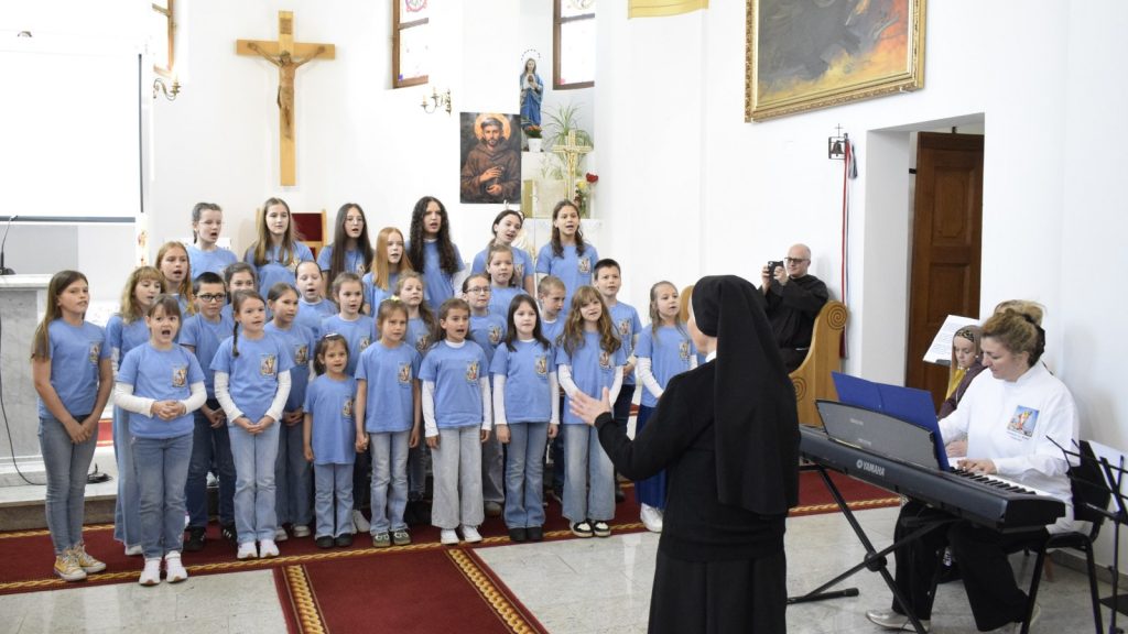 A group of children in matching blue shirts singing in a church, led by a nun conducting at the front while a pianist accompanies on a keyboard nearby.