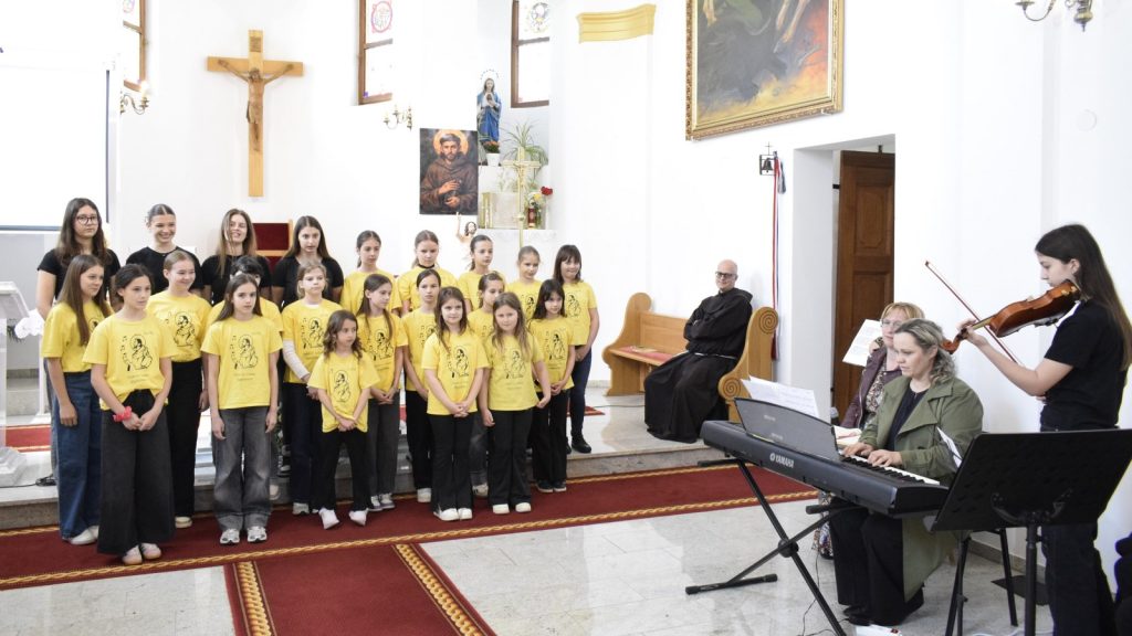 Children's choir wearing yellow shirts performing in a church, with musicians and a monk seated in the background.