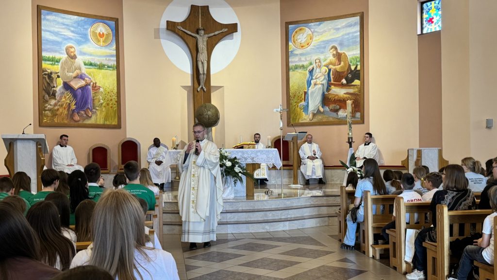 Priest in white vestments speaks at the altar during a church service with parishioners listening in pews behind.