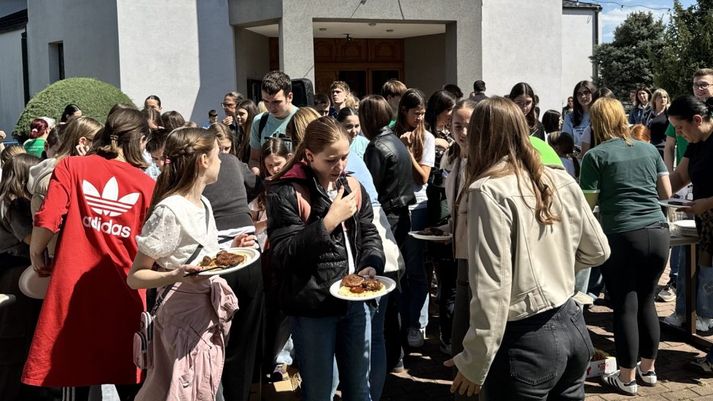 Crowd of teens and young adults outside a building, many holding plates with pastries at a sunny outdoor event.
