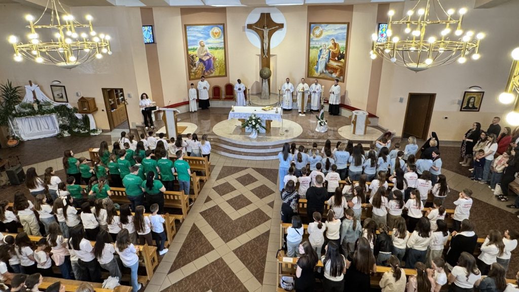 Wide view of a church service: altar with crucifix and priests in white vestments, a lector at a podium, and a large congregation of children and adults seated and standing all around.