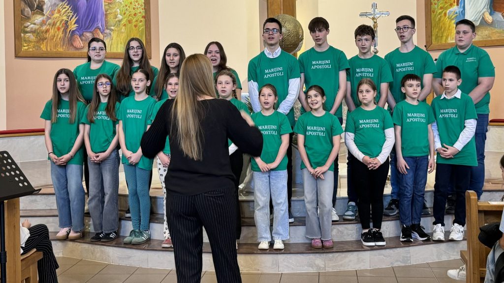 Group of children in green shirts singing while a conductor leads them in a church setting on stage steps with paintings and a crucifix in the background.