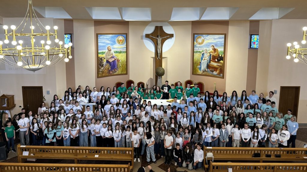 Large group of children and adults posing for a group photo in a church, with a central crucifix and two paintings behind them.