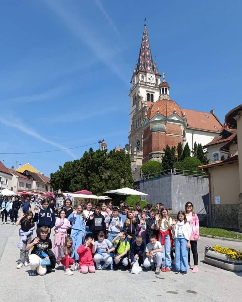 Group of smiling children posing for a photo in a town square with a tall ornate church tower behind them in clear blue sky.",