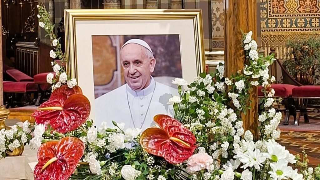Framed portrait of Pope Francis surrounded by white flowers and red anthuriums in a church setting.