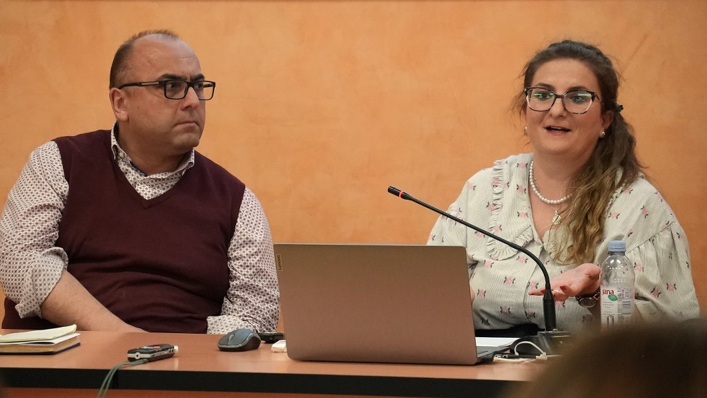 Two panelists at a conference table: a man in a burgundy vest and glasses on the left, and a woman with glasses speaking into a microphone beside a laptop and water bottle.