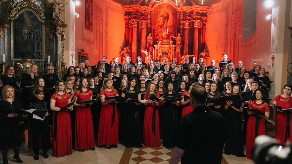 Large choir in red and black gowns performs in a grand church with an ornate altar behind them; conductor seen from the back in the foreground.