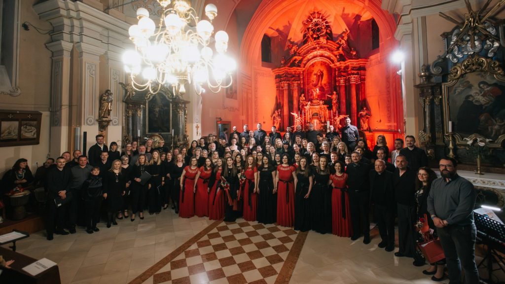 Large choir posing in a church with a red-lit altar in the background; singers wear black and red attire.