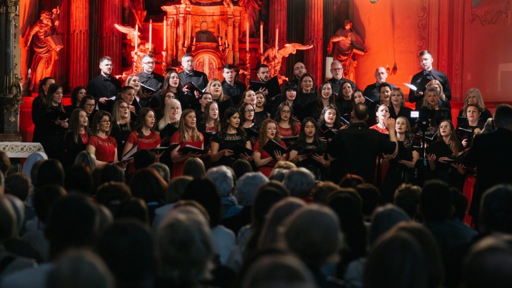 A large choir in red and black performs on a church stage while a conductor leads from the front, with an audience watching in the foreground.
