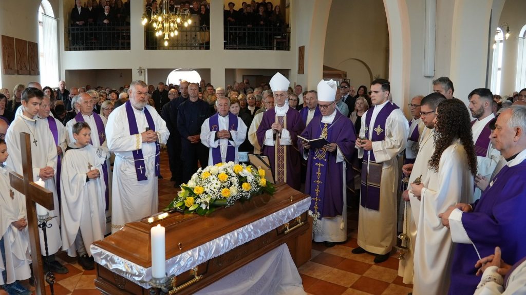 Catholic funeral with priests in purple vestments around a wooden casket adorned with yellow and white flowers, candles lit, in a crowded church hall.