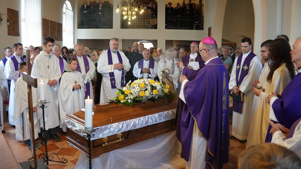 Catholic funeral in a church: priests in purple vestments gather around a flower-decorated coffin as mourners look on.