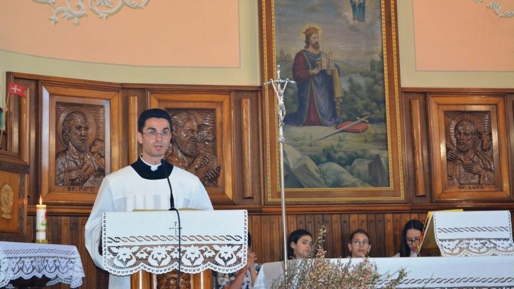 Priest in white liturgical robes speaks from a decorated church pulpit, with wooden panels and a large framed painting behind him.
