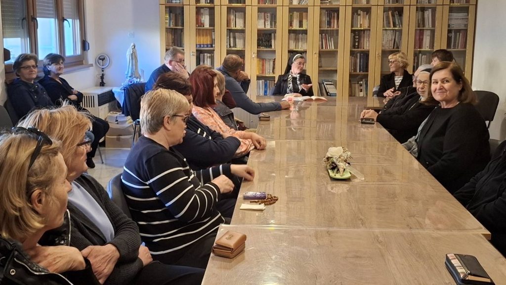 Group of adults seated along a long wooden table in a library, with a nun speaking at the head of the table.