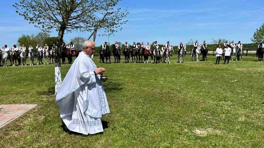Priest in white vestments walks across a grassy field, holding a censer, while a line of riders on horseback stands behind under a blue sky.
