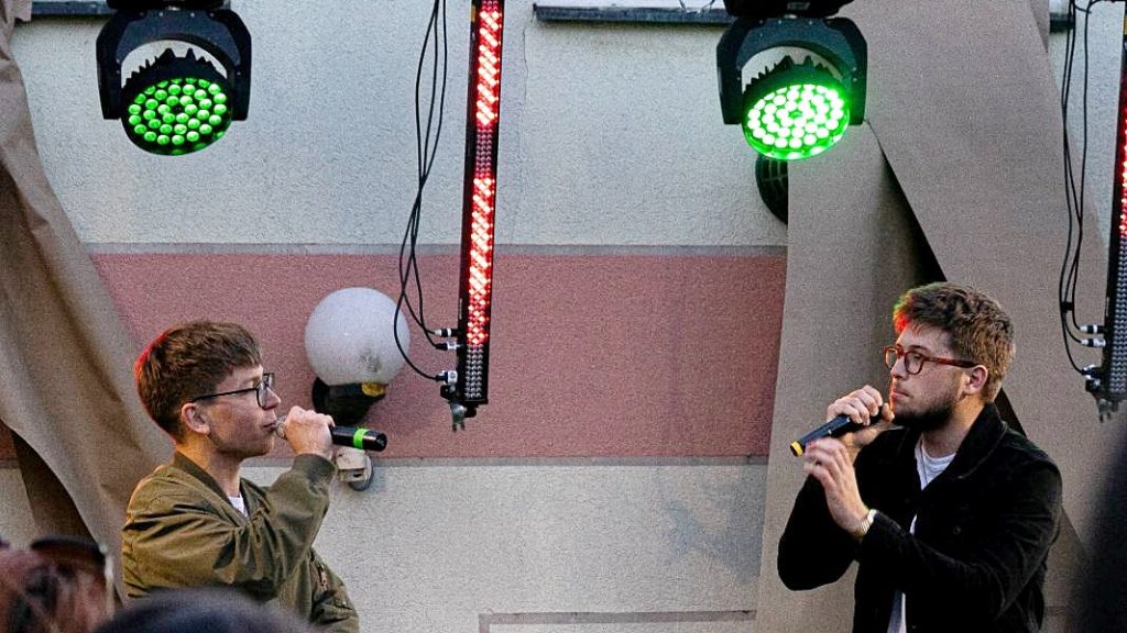 Two male singers wearing glasses perform on an outdoor stage, each holding a microphone, with green and red stage lights above.