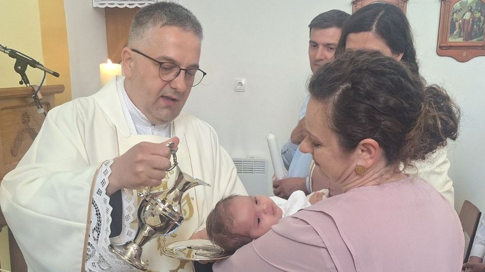 Priest in white robes pours holy water from a silver pitcher as a mother holds her newborn during a baptism-like ceremony.