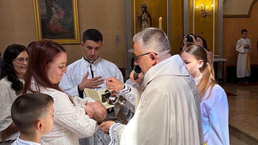 Priest pours baptism water over a baby held by the mother during a church ceremony, surrounded by family in white.