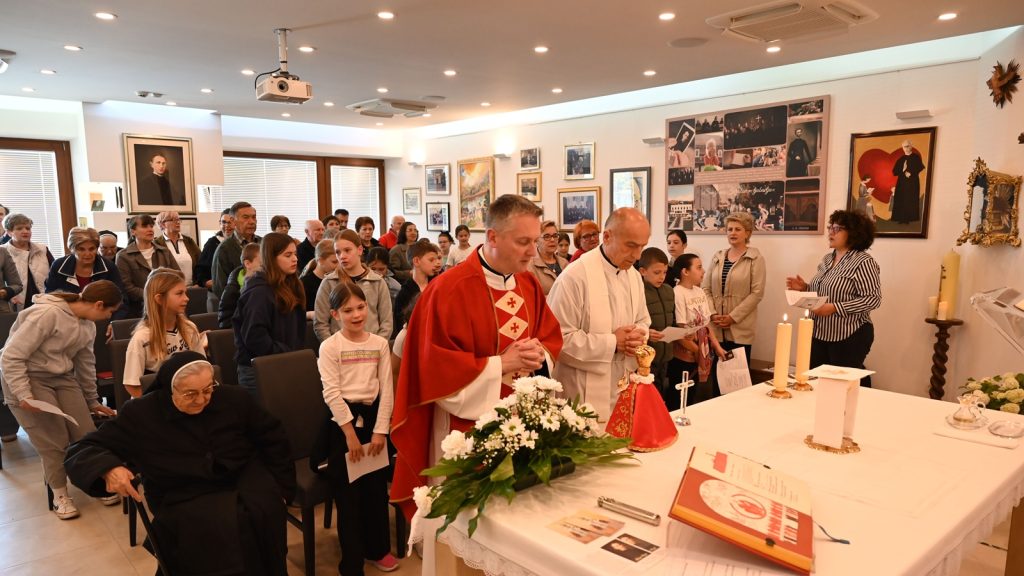 Priest in red vestments leading a Mass at an altar with flowers, as children and adults stand in prayer behind him.