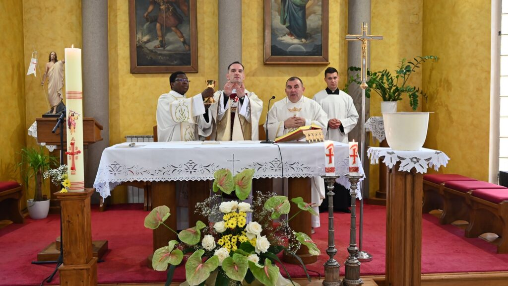 Priest elevating the Eucharist during Mass at an ornate church altar, flanked by two priests and a server, with candles and flowers nearby.