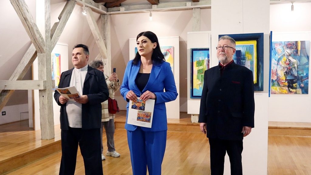 Three adults stand in a contemporary art gallery; a woman in a blue suit holds a program while two men flank her, with colorful paintings in the background.