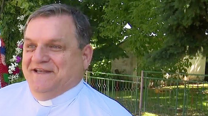 Smiling priest wearing a white clerical shirt outdoors, with greenery and a fence in the background.