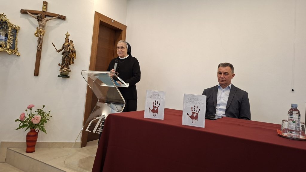 A nun speaks at a clear podium with a microphone, standing beside a seated man in a suit at a table with booklets and water bottle in a religious room.