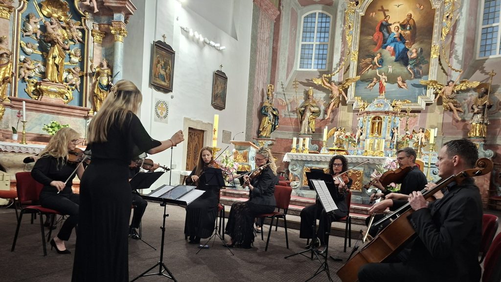 A string ensemble performs in a richly decorated church with gilded statues and a large religious painting above the altar.