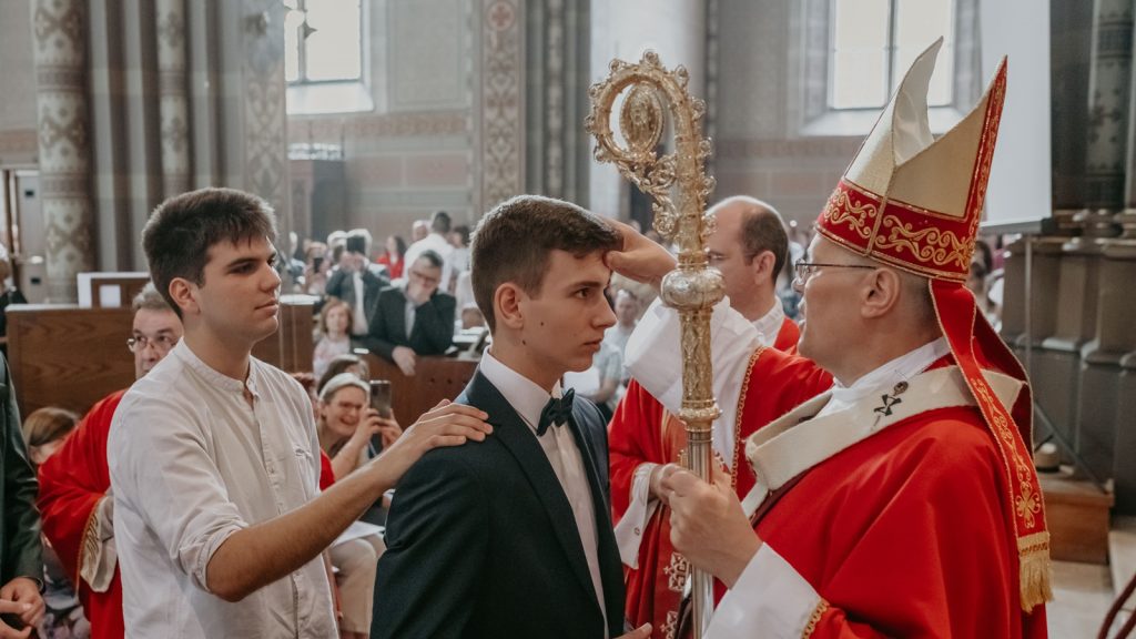 A clergyman in red ceremonial robes blesses a young man in a dark suit with a hand on his head during a church ceremony.