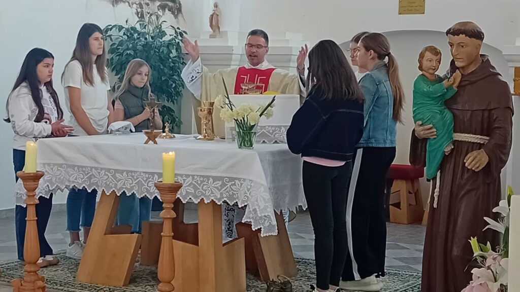 Priest in white vestments blesses a group of girls at an altar during a Catholic service, with candles and chalices on the table.