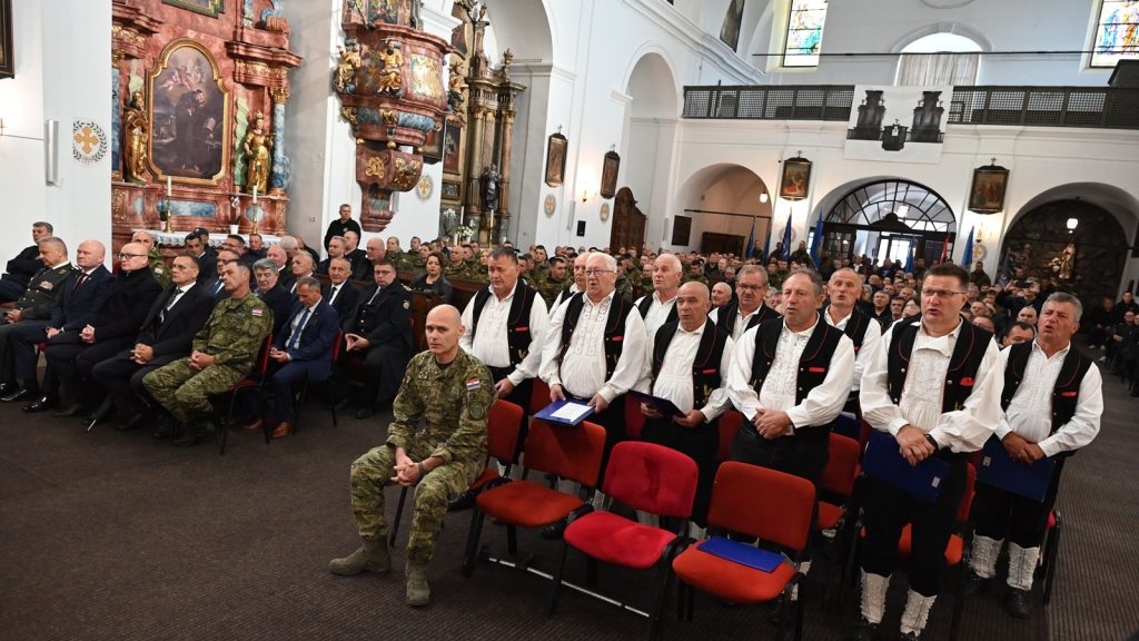 A formal ceremony inside a church with uniformed soldiers and men in suits seated in red chairs near an ornate gilded altar on the left.