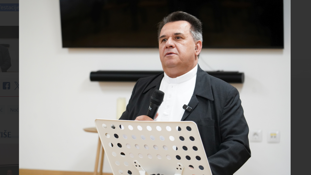 Clergyman in a black suit and white clerical collar speaks into a handheld microphone at a perforated white lectern.