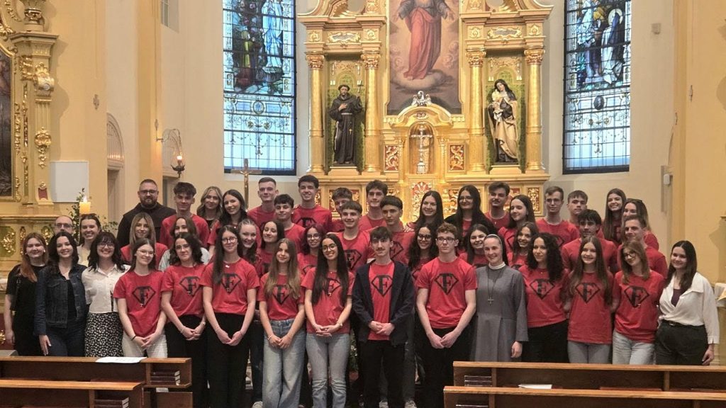 Group of students in red shirts posing for a group photo inside a church with an ornate gilded altar in the background, and a few adults at the edges.