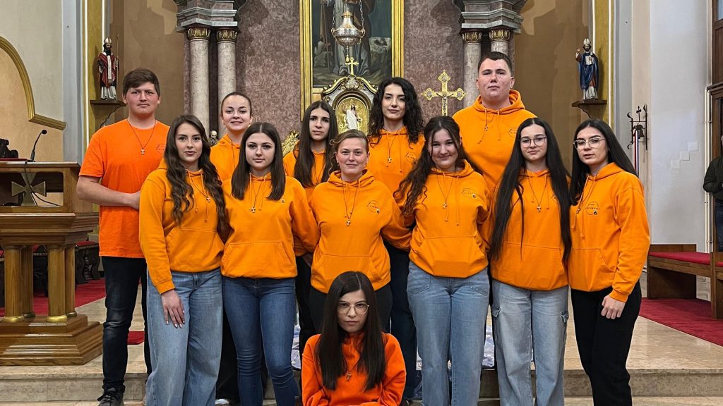 Group of fourteen teenagers and one young adult in matching orange hoodies posing on church steps in front of an ornate altar with gold accents and religious statues.