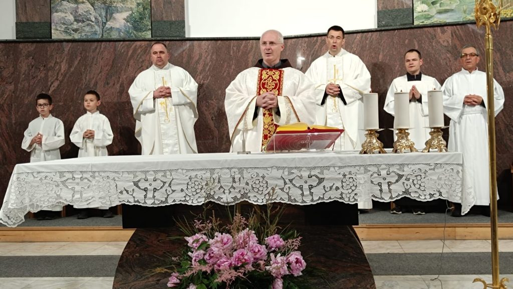 Catholic altar service with a central priest in ornate red stole, flanked by priests and two altar boys, behind a lace-clothed altar with candles and a Bible