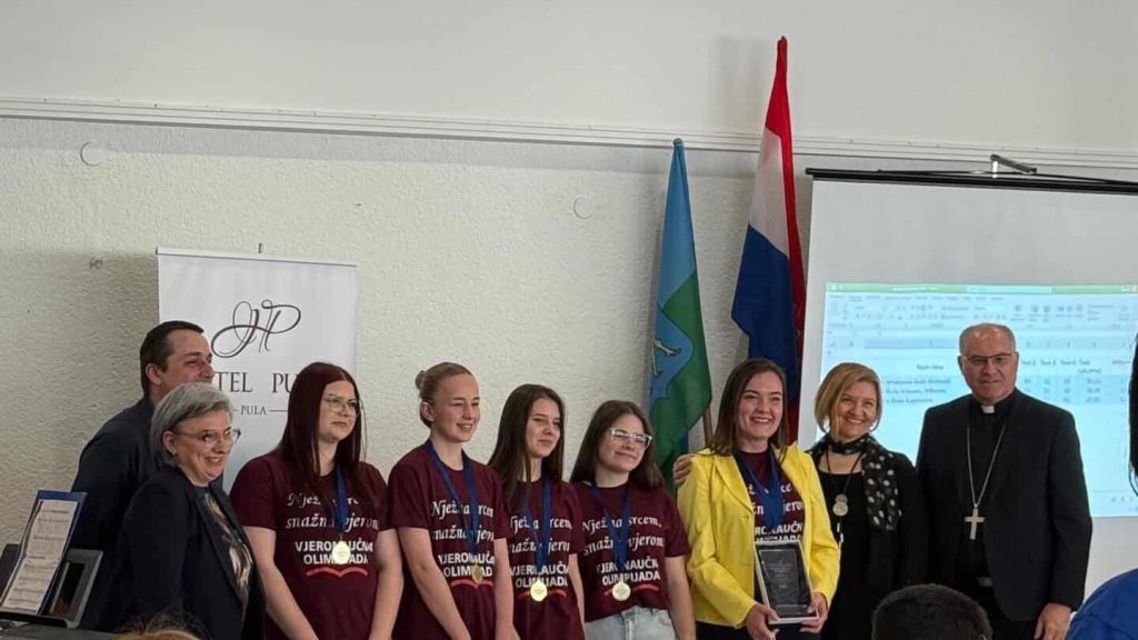 Group of students and adults posing for a ceremony photo, some wearing maroon shirts and medals, with a trophy/podium nearby and flags in the background.