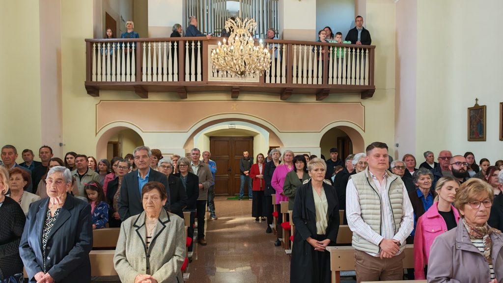 Crowded church interior with people standing in pews during a service, a balcony with a chandelier above.