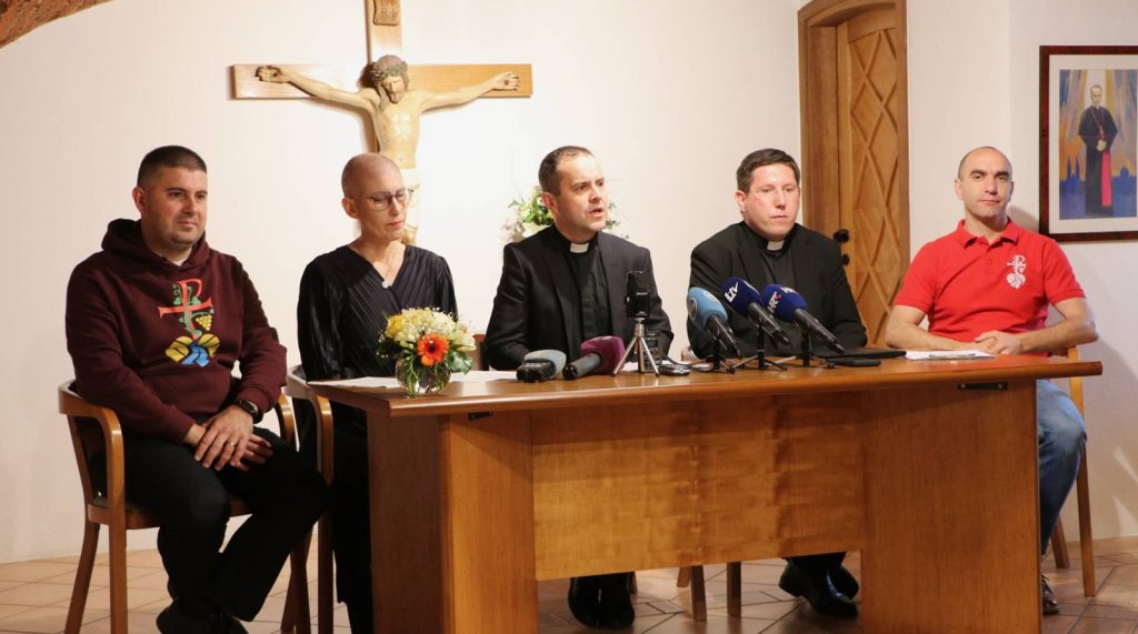 Group of five people at a press conference table; multiple microphones in front, crucifix on the wall behind them.