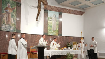 Catholic Mass in progress with priests at the altar and altar servers assisting in a church setting, candles lit and a white cloth-draped altar.
