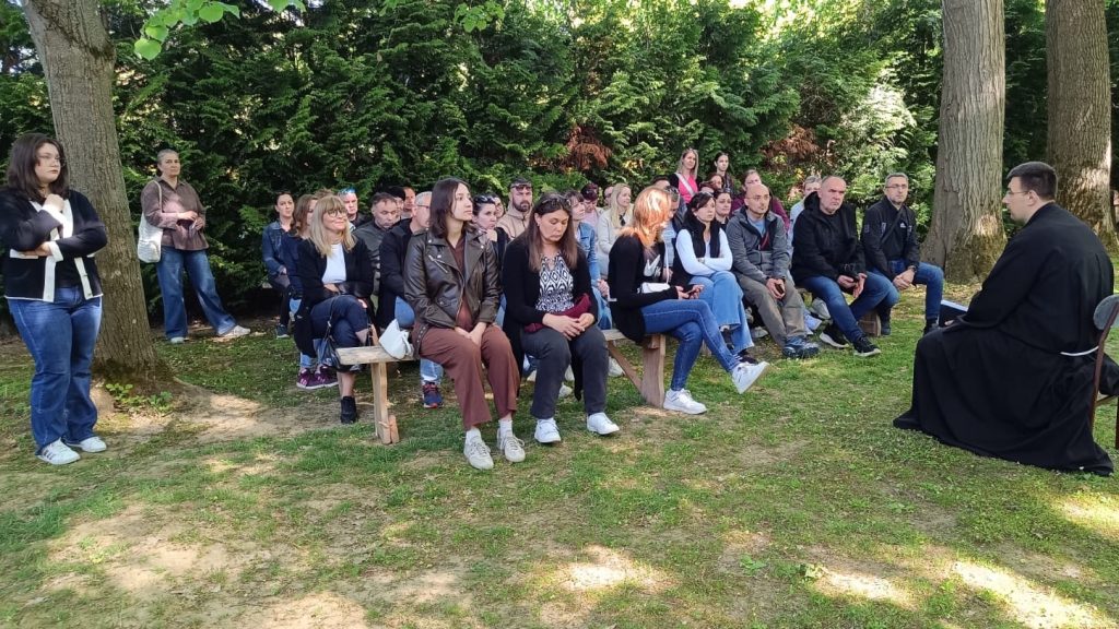 Outdoor group gathering: a priest speaks to a crowd seated on benches in a park with trees in the background.