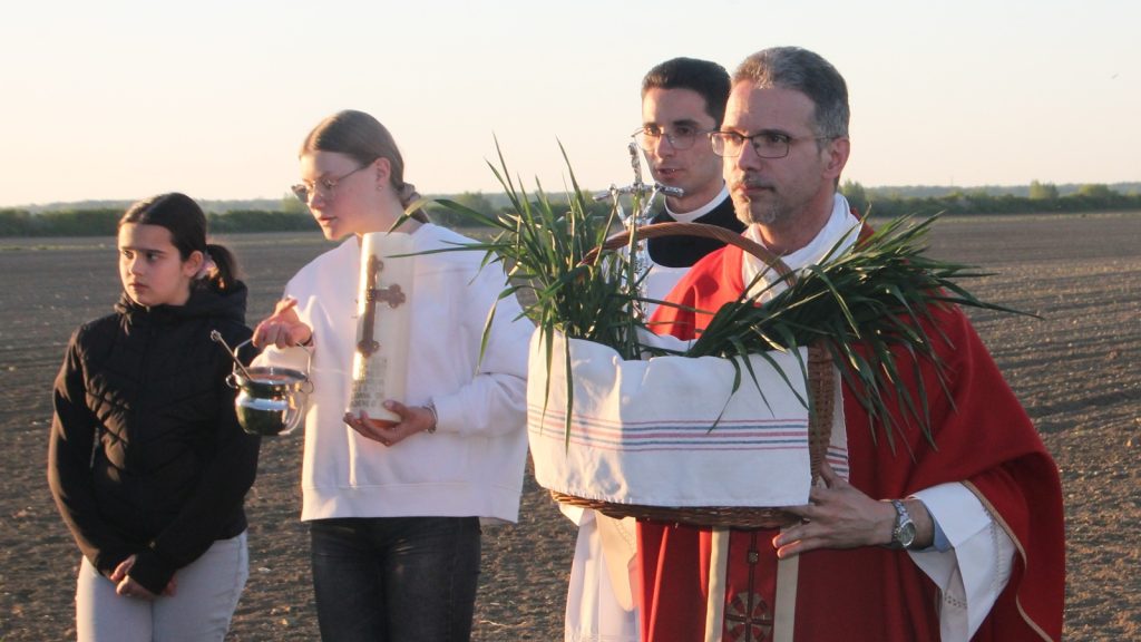 Outdoor religious procession at sunset: a priest in red vestments carries a basket of palm leaves, accompanied by assistants and a young girl in a black jacket.