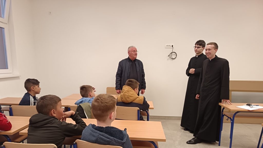 Older man in a dark jacket stands at the front of a classroom as two priests in black cassocks talk to him, while students sit at desks watching.